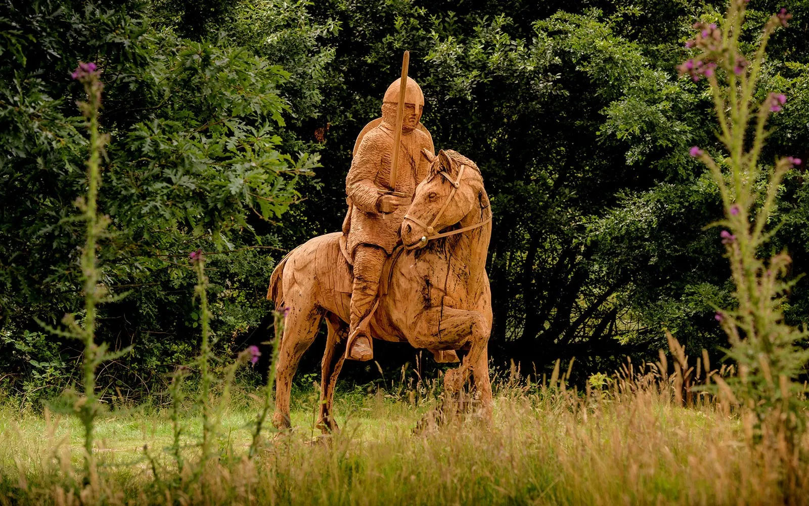 A carved wooden statue of a mounted Norman knight on the battlefield at Battle, among thistles and long grass