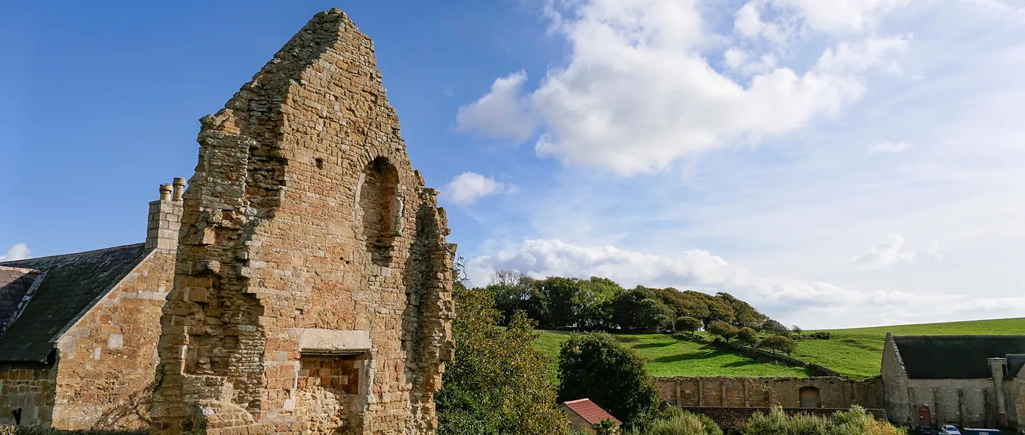 The tall ruined gable of Abbotsbury Abbey in warm golden stone, beside a cottage, with the long tithe barn and a green hillside beyond under a blue sky