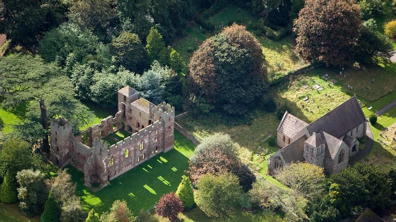 Aerial view of the roofless red sandstone shell of Acton Burnell Castle on a sunny lawn, beside the parish church among trees