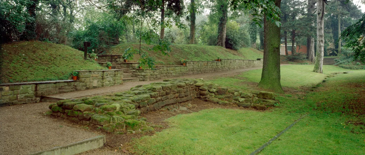 Moss-covered Roman wall foundations beside a gravel path and terraced stone walls among trees at Aldborough