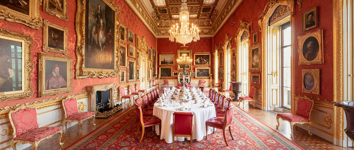 The Waterloo Gallery laid with a long dining table beneath red damask walls hung with paintings