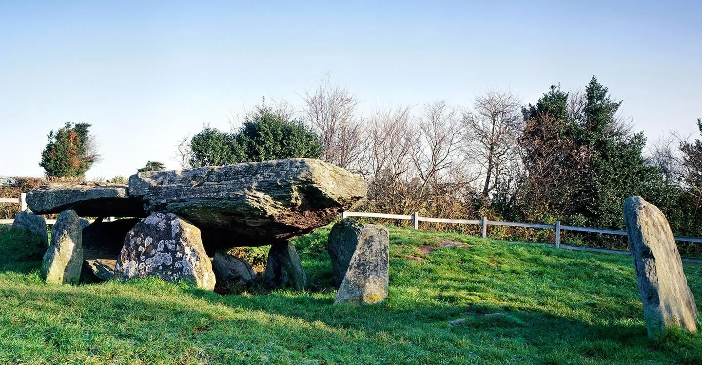 The huge capstone of Arthur's Stone resting on upright stones on a grassy mound, with hedges and a wooden fence under a clear sky