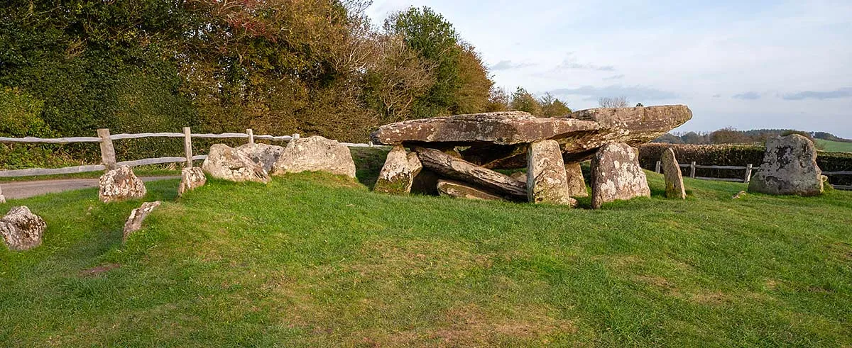 The Neolithic chamber tomb of Arthur's Stone and its outlying stones on a lawn beside a lane, with hedges and fields in autumn light