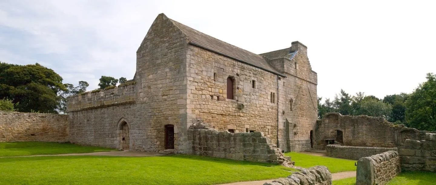 The gabled hall block and ruined outer walls of Aydon Castle around a grassy courtyard, with trees beyond on a summer day