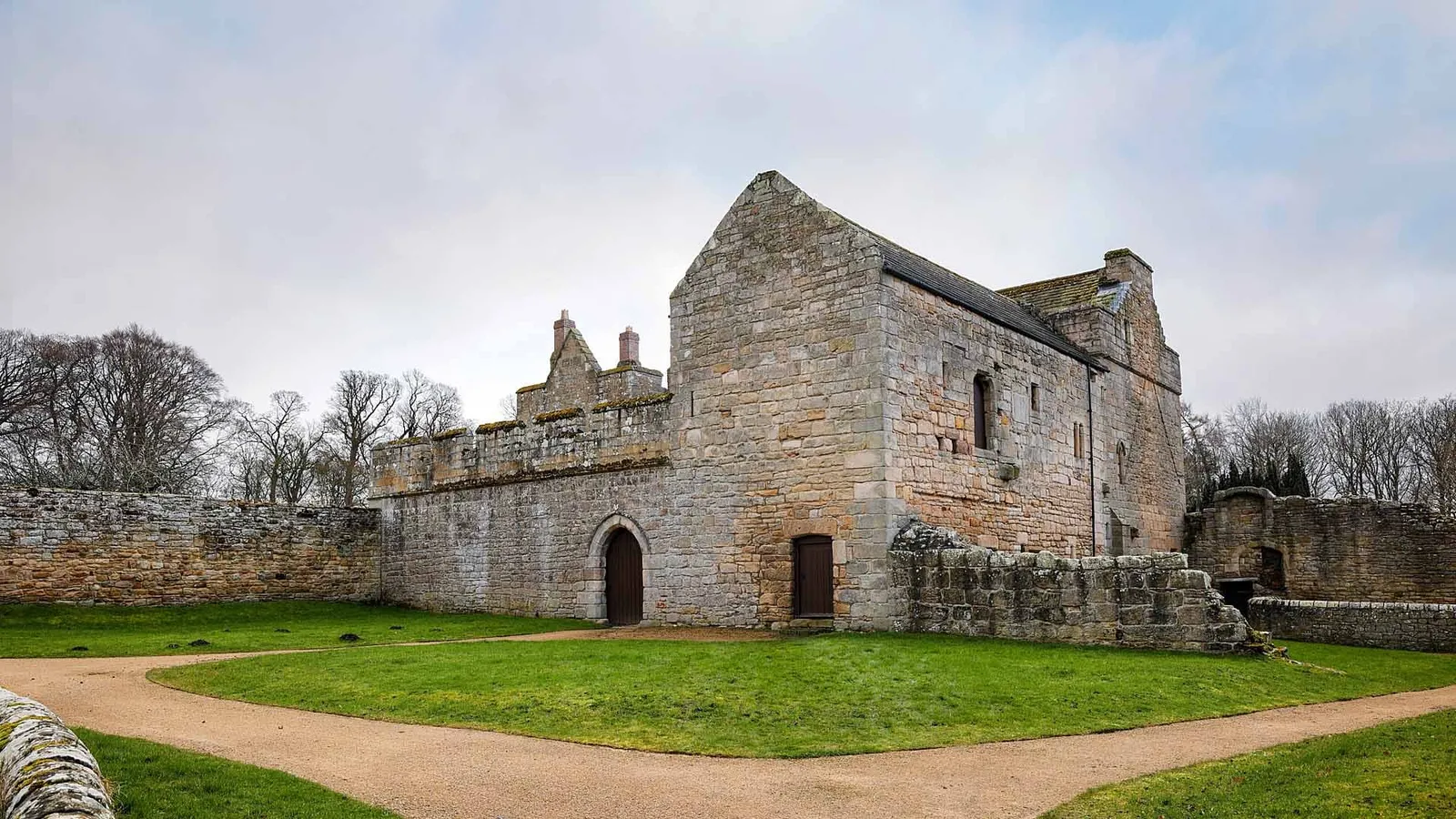 The tall stone hall range of Aydon Castle with its crenellated curtain wall and an arched doorway, beside a curving gravel path and lawn