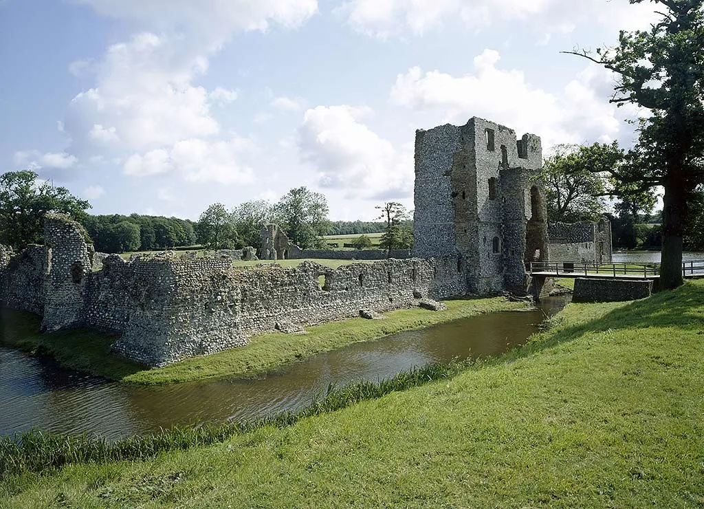 The ruined flint inner gatehouse and curtain wall of Baconsthorpe Castle beside its moat and timber bridge, under summer clouds