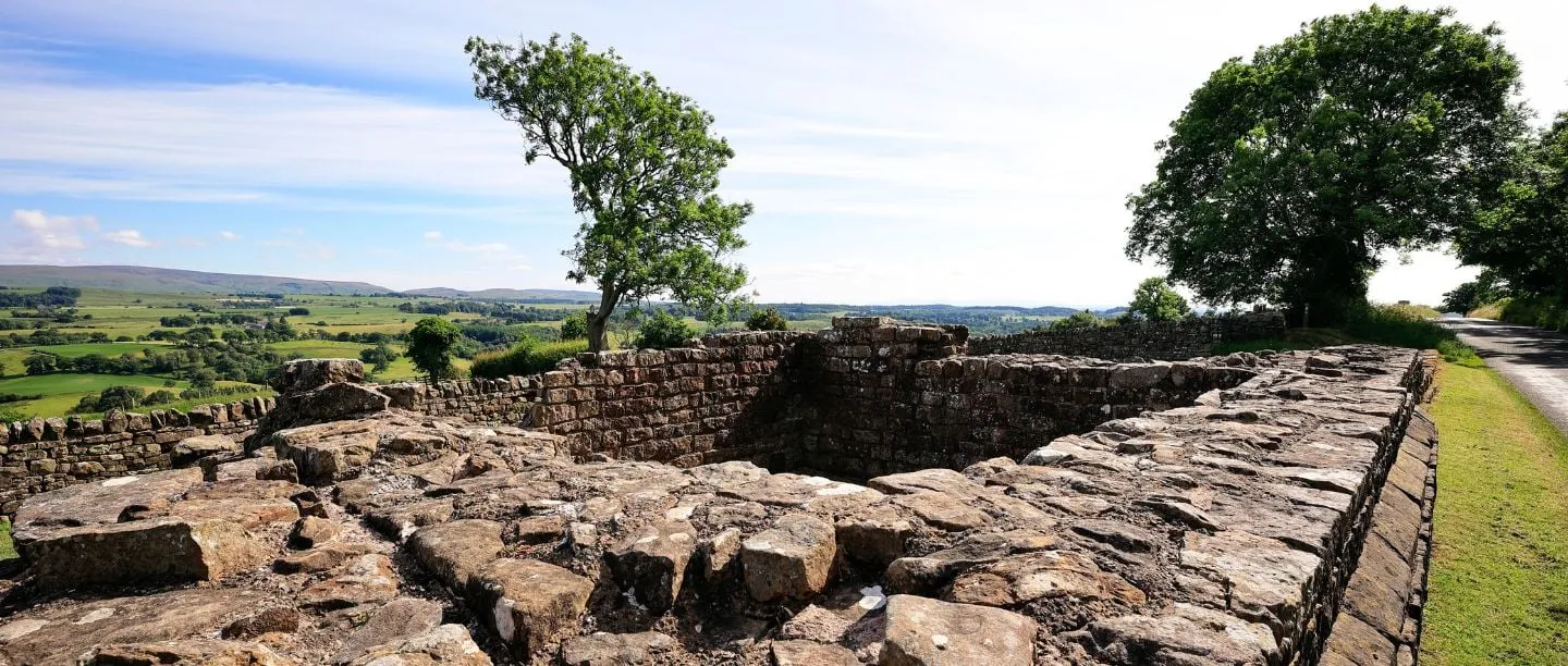 The stone footings of Banks East Turret on Hadrian's Wall beside a lane, with trees and wide views over green farmland to distant hills