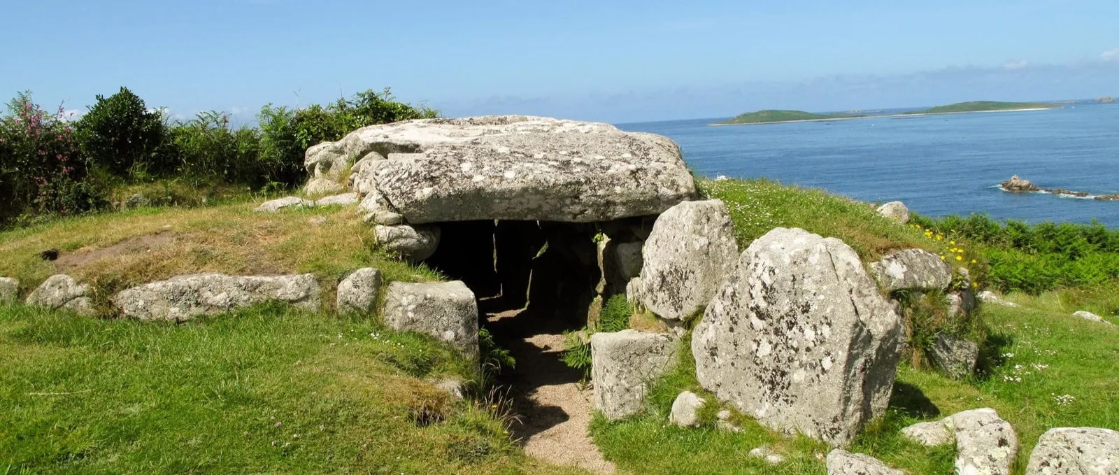 Bant's Carn entrance grave, a massive lichen-covered capstone over a dark passage, with the blue sea and islands beyond