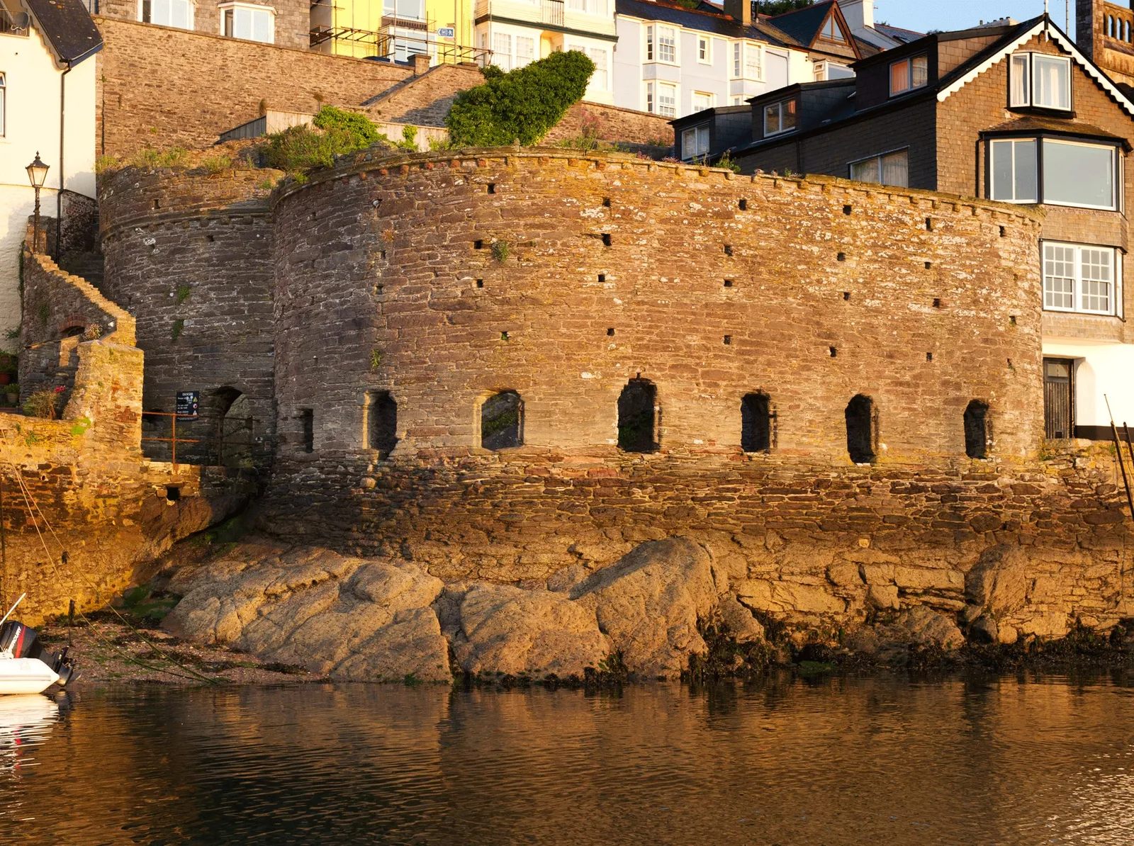 The curved stone gun wall of Bayard's Cove Fort with a row of arched gunports, on rocks at the water's edge in golden evening light, with houses above
