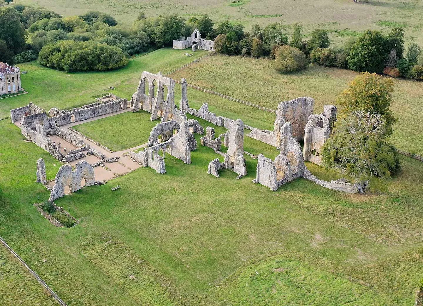 An aerial view of Bayham Old Abbey's roofless church, cloister and outbuildings on mown grass, with the Dower House and gatehouse beside woodland