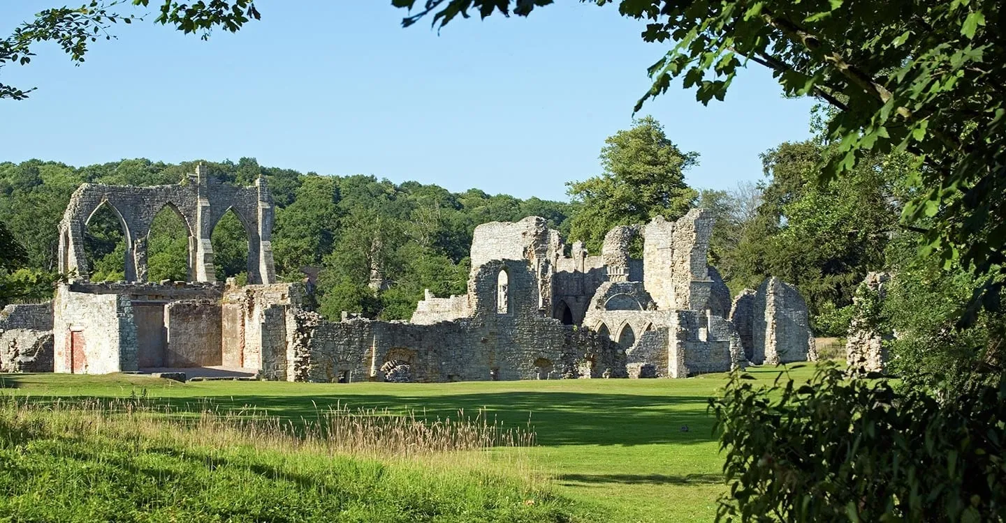 The ruined church and chapter house of Bayham Old Abbey across a sunlit lawn, framed by overhanging trees under a clear blue sky