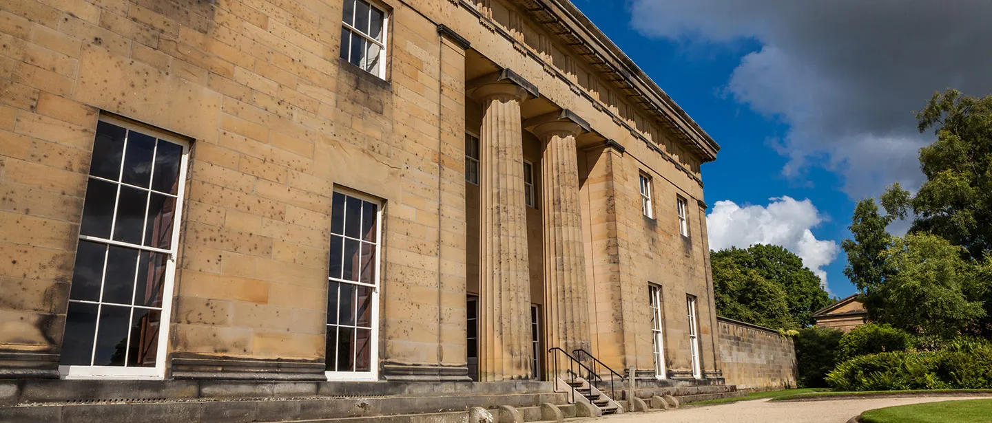The Greek Doric columns and honey-coloured sandstone of Belsay Hall under a stormy blue sky
