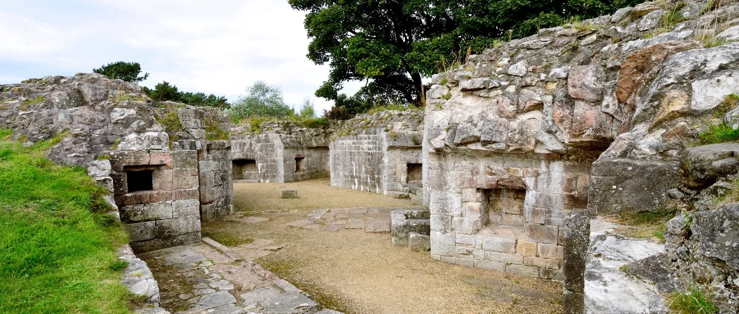 Ruined stone chambers and window openings of Berwick Castle set into rock, with a gravel floor and trees above