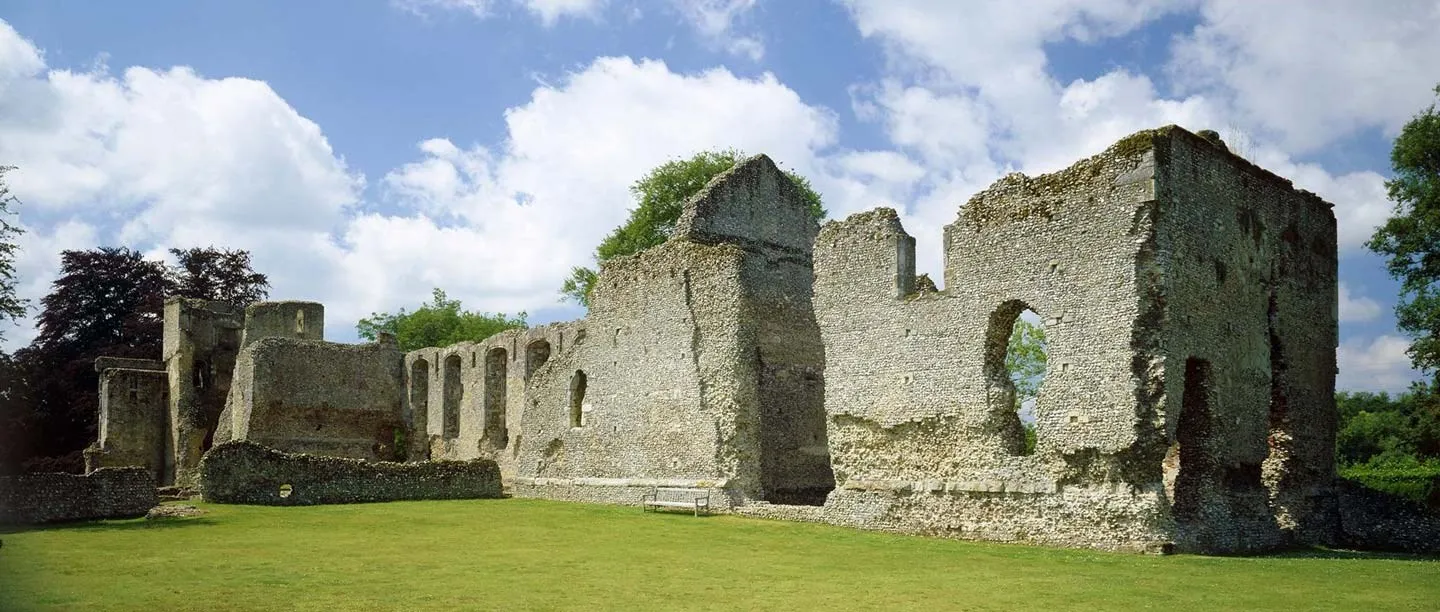 The tall flint walls and empty window arches of the great hall and west range at Bishop's Waltham Palace, beside a lawn and bench under a cloudy blue sky