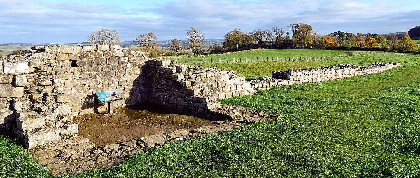 Black Carts Turret on Hadrian's Wall, its square stone walls and an interpretation panel, with the wall running on across autumn fields