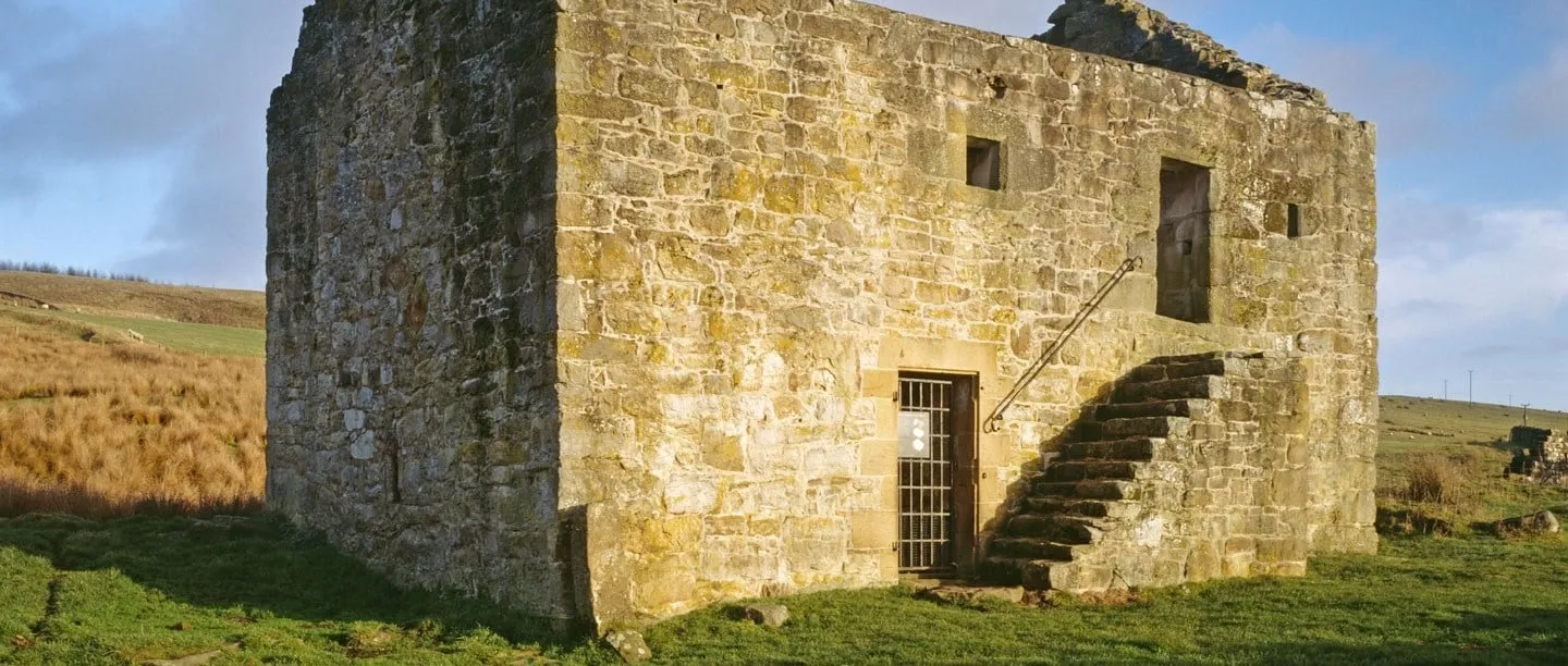 Black Middens Bastle House, a roofless two-storey stone farmhouse with an outside stone stair and barred door, on moorland grazing under a blue sky