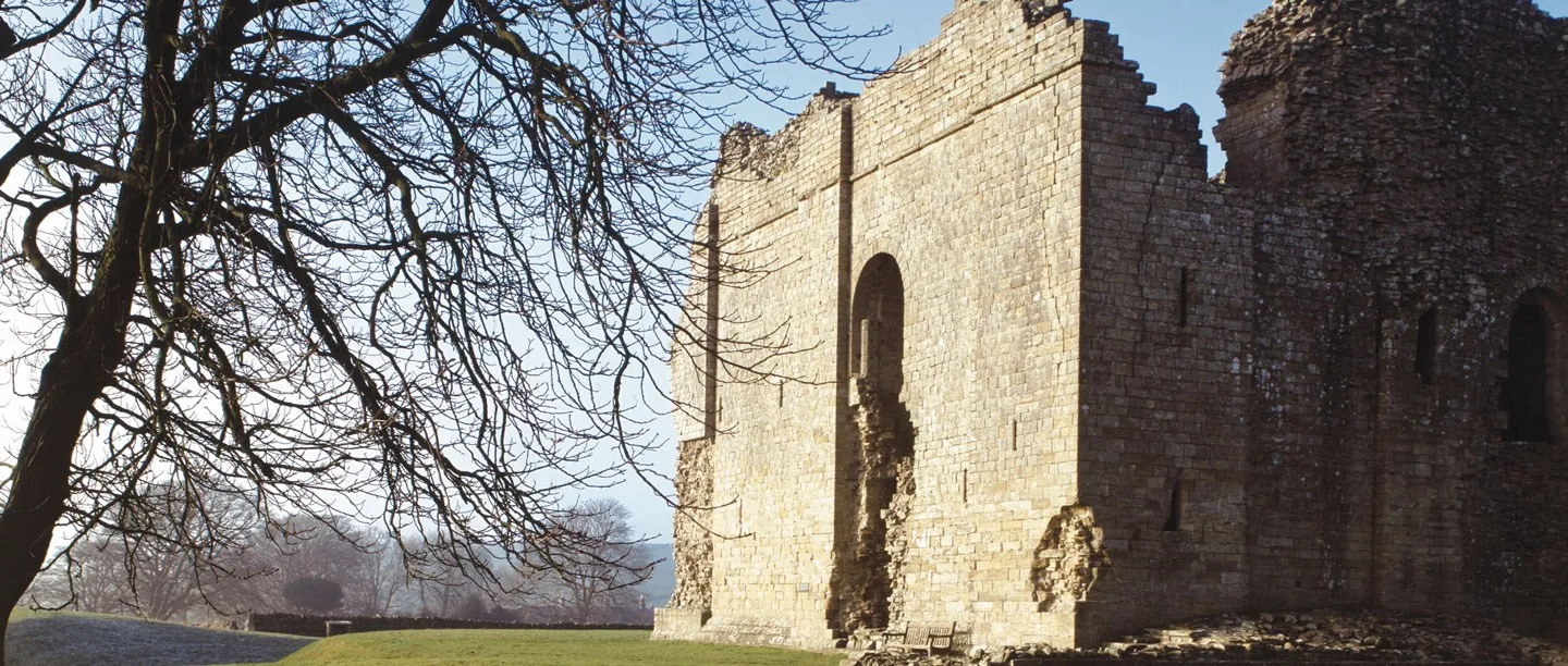 The tall roofless keep of Bowes Castle with a round-arched window opening, beside a bare tree and a frosty lawn in winter sun