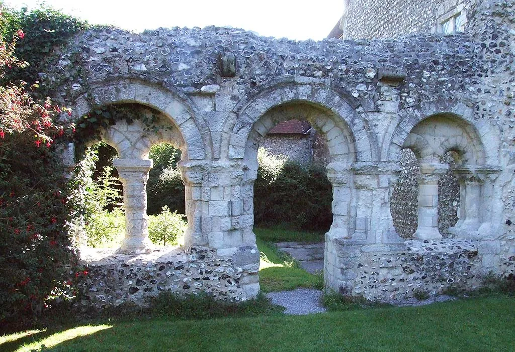 Round Norman arches on stout columns in the flint wall of the Boxgrove Priory chapter house, with a lawn, gravel path and fuchsia