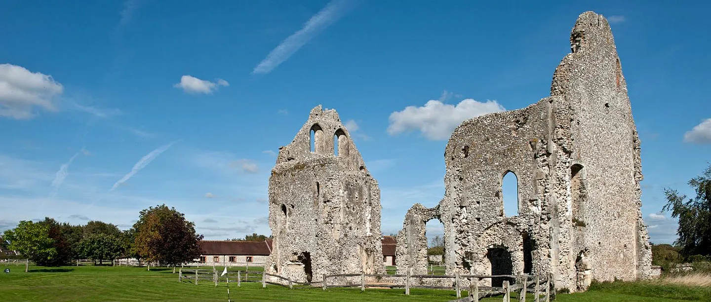 The roofless flint guest house of Boxgrove Priory rising from a wide lawn with a post-and-rail fence, beside farm buildings and trees