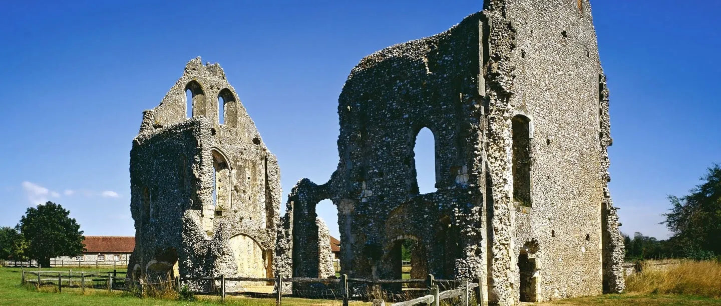 The tall flint ruins of the guest house at Boxgrove Priory with empty pointed windows, behind a wooden fence under a deep blue sky