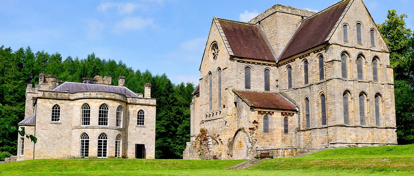 The Norman church of Brinkburn Priory and the neighbouring manor house on a green lawn below dense conifer woods, under a blue sky