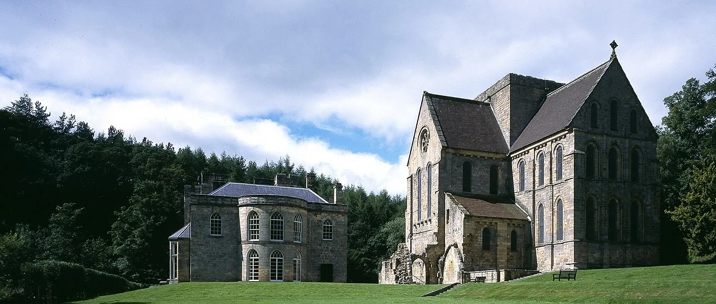 Brinkburn Priory church and the manor house across a lawn, beneath billowing clouds and dark woodland