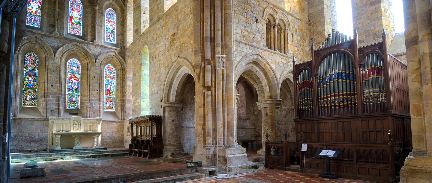 Inside Brinkburn Priory church, pointed arches beside a painted organ, with an altar and colourful stained glass windows
