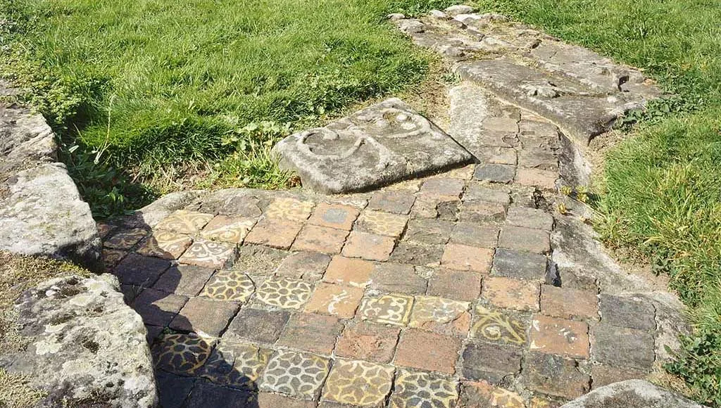 Patterned medieval floor tiles in yellow and brown set into the ground of Buildwas Abbey's chapel, beside worn stone slabs and grass