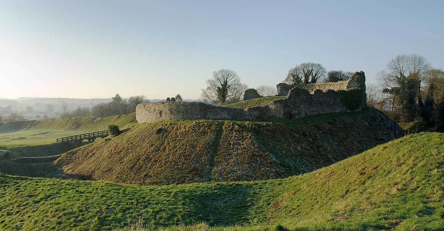 The ruined flint walls of Castle Acre's keep on its high earthwork mound above a grassy ditch and wooden bridge