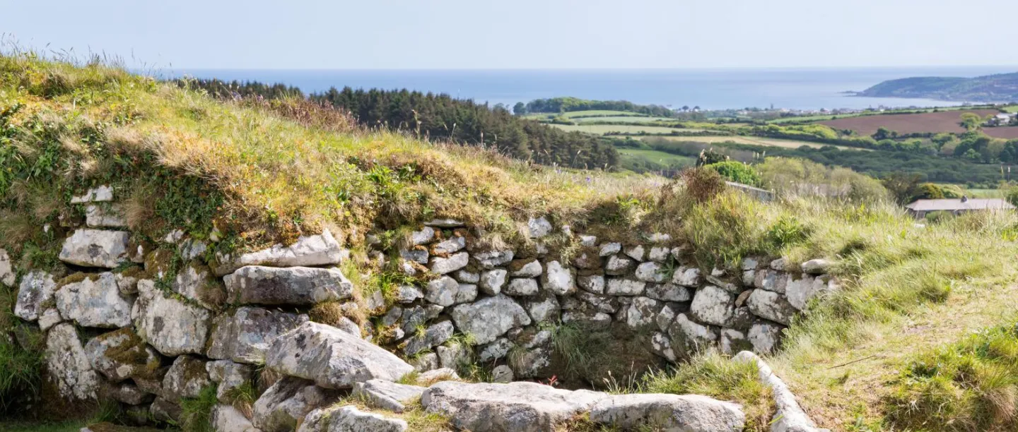 The dry-stone wall of a Chysauster house in long grass, looking over fields and woods to the sea at Mount's Bay