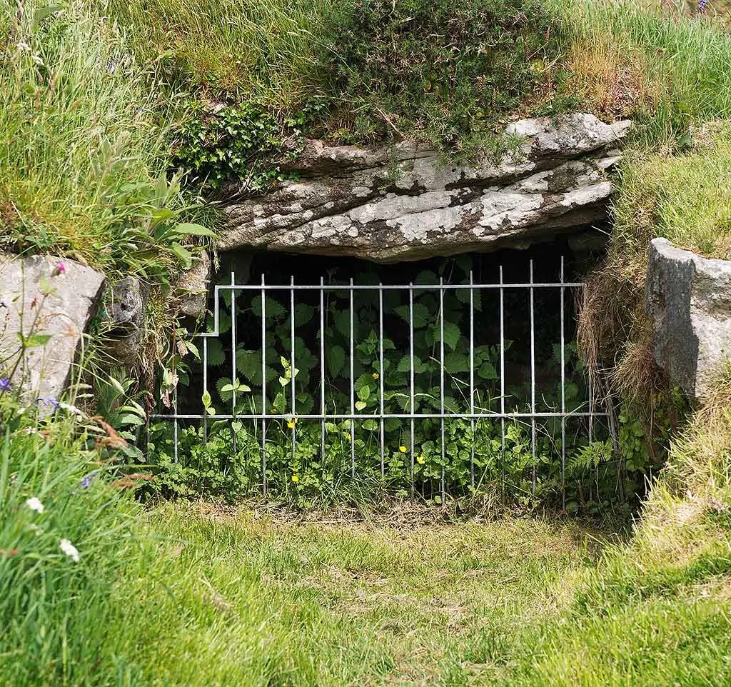 The low entrance of the fogou at Chysauster, an underground passage beneath a granite lintel, behind an iron grille among nettles