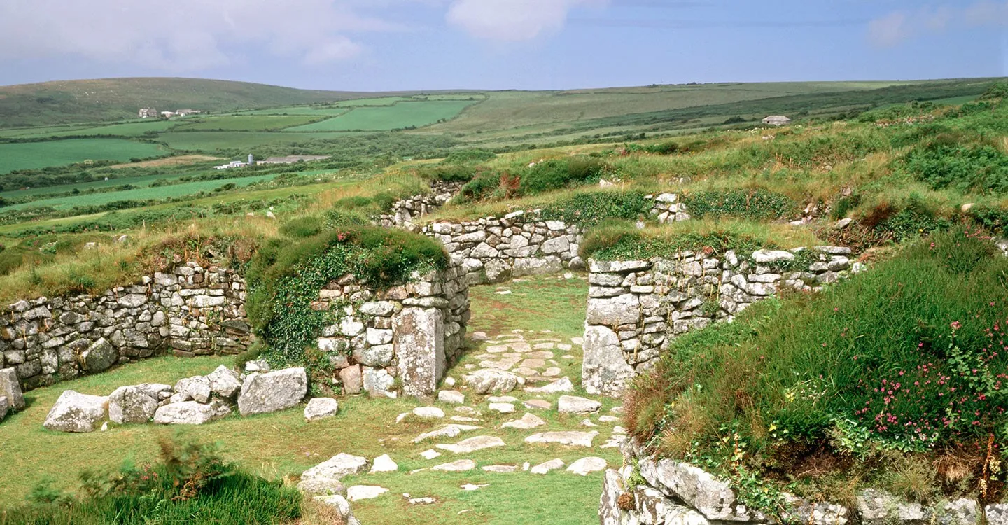 The stone-walled courtyard houses of Chysauster Iron Age village with a paved entrance passage, among heather and grass in rolling Cornish farmland