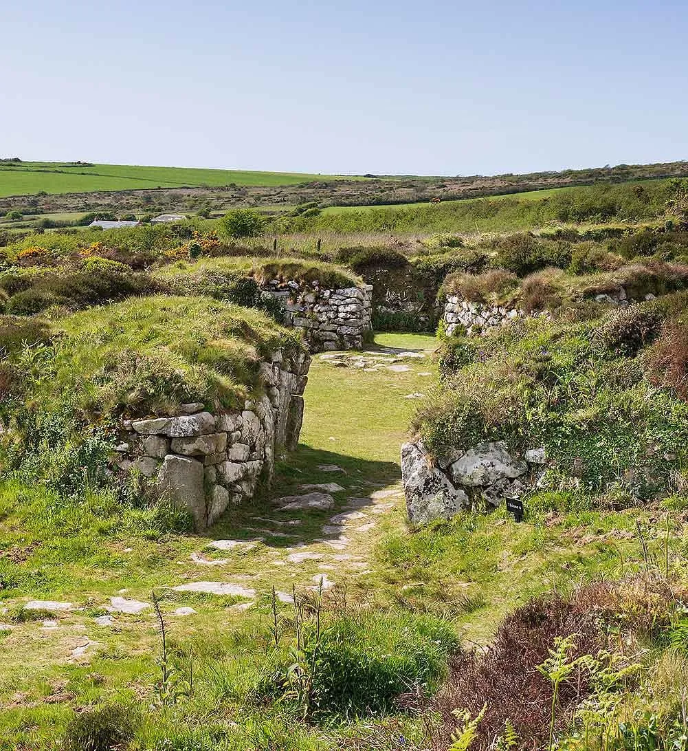 A grassy passage between curving dry-stone walls into house 4 at Chysauster, with paving stones, gorse and green fields beyond