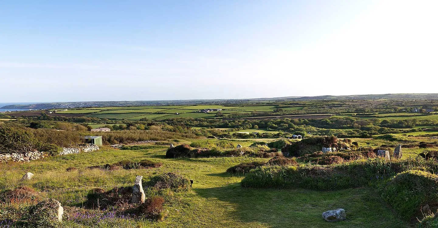 The low grassy remains of Chysauster village in evening light, with standing stones and rolling fields stretching to the sea