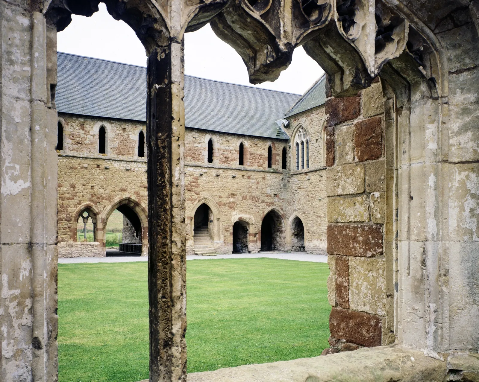 The cloister lawn and dormitory range of Cleeve Abbey seen through the carved stone tracery of a cloister window