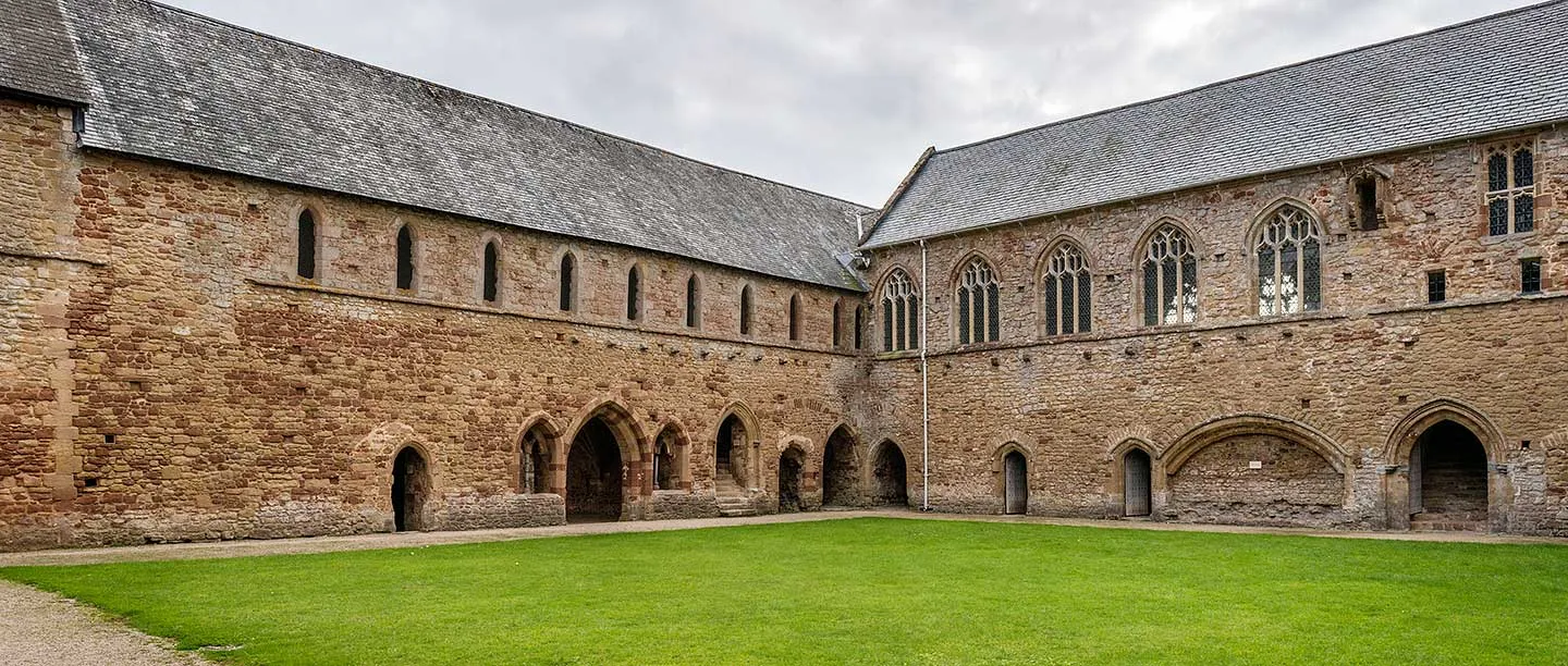 The two-storey dormitory and refectory ranges of Cleeve Abbey around the grassy cloister, built of red and buff sandstone under slate roofs
