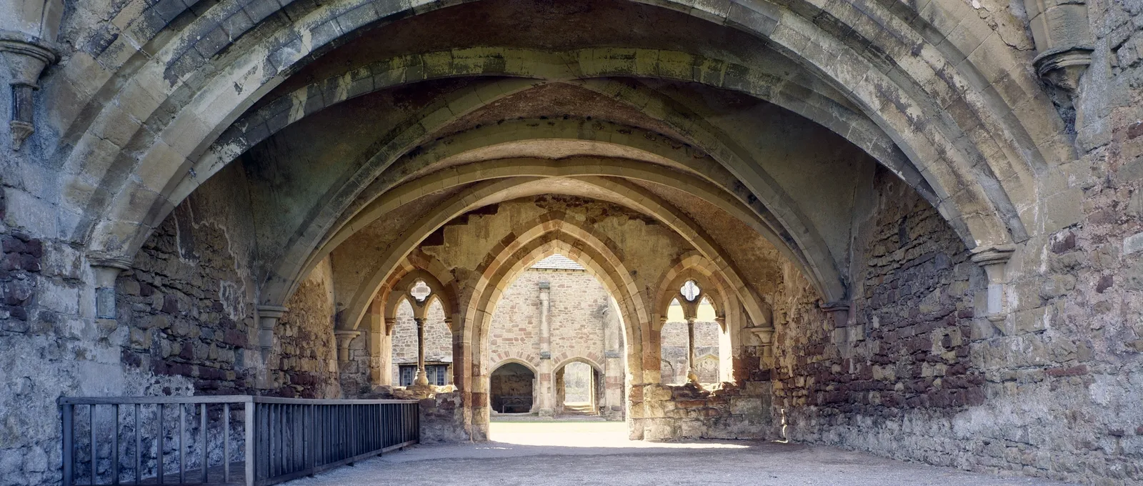 Pointed arches of the chapter house entrance at Cleeve Abbey framing the ruined church beyond, on a gravel floor in soft sunlight