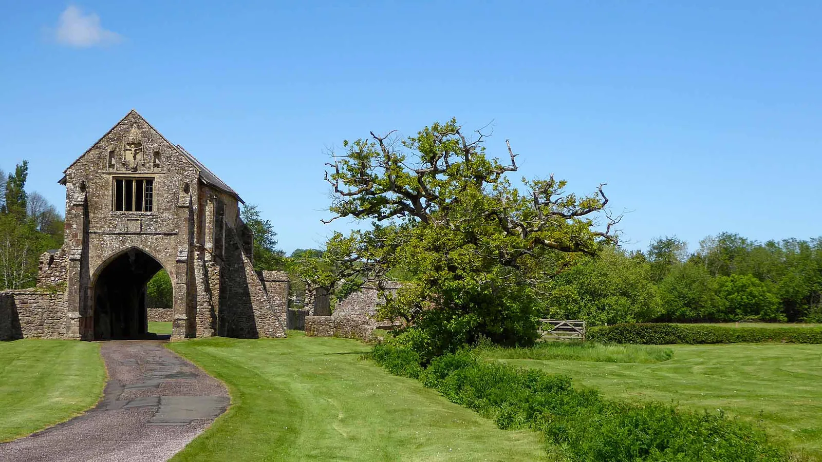 The medieval gatehouse of Cleeve Abbey with a carved niche above its tall pointed archway, a path leading through and an oak tree on a sunny day