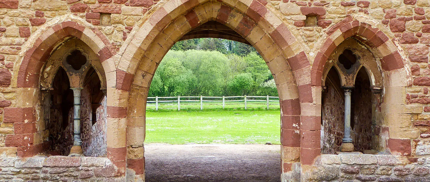 Banded red and cream sandstone arches of the chapter house entrance at Cleeve Abbey, with a lawn and fence beyond