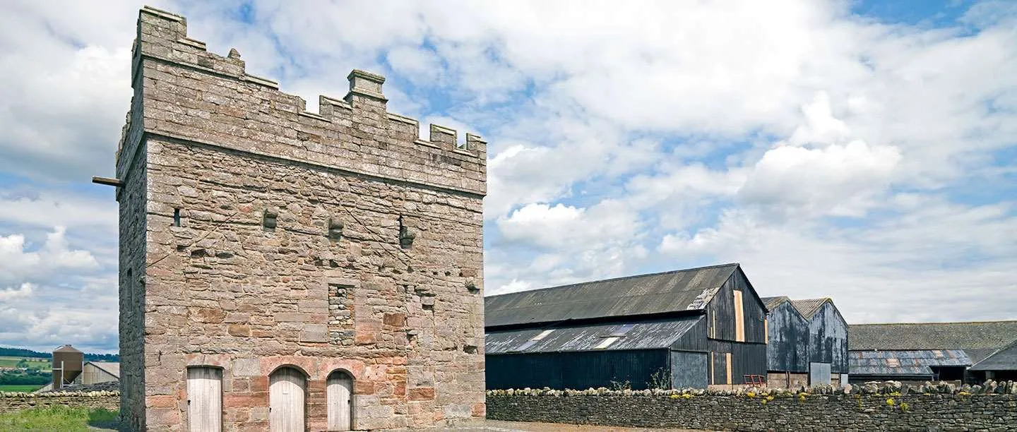 Clifton Hall, a battlemented sandstone tower with three arched doors, beside a dry-stone wall and black farm barns