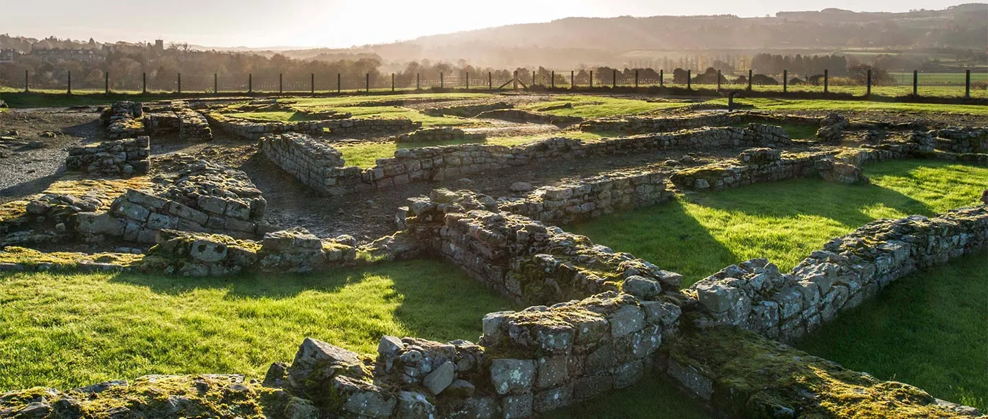 Low walls of Roman Corbridge's excavated buildings in hazy morning sun, with the Tyne valley beyond