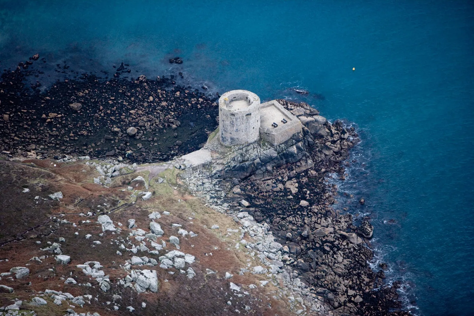 Cromwell's Castle from the air, the round tower and gun platform with two cannon on rocks at the edge of a turquoise sea
