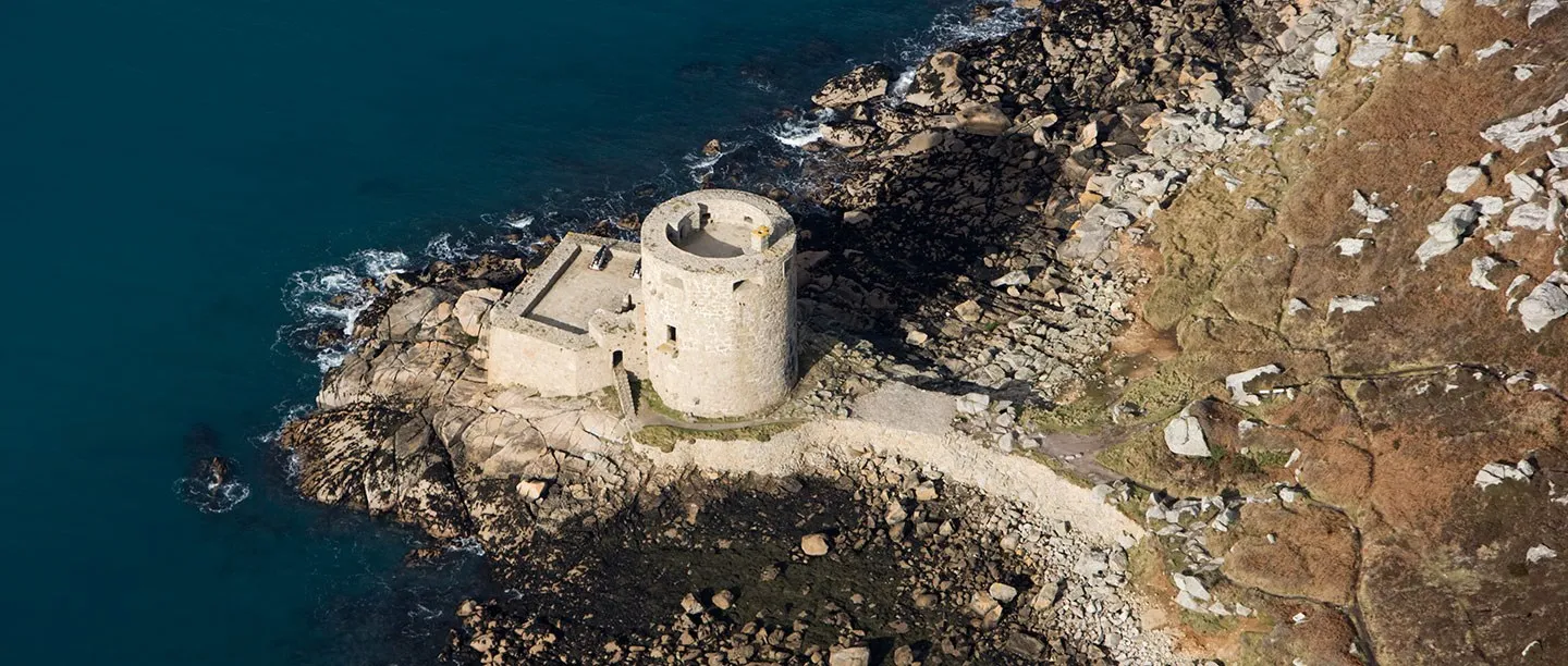 An aerial view of Cromwell's Castle, a round granite tower with a square gun platform on a rocky point above dark blue sea