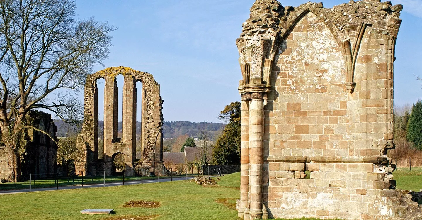 The soaring lancet windows of Croxden Abbey's ruined south transept, with a carved column in the foreground, lawns and a lane under a blue sky