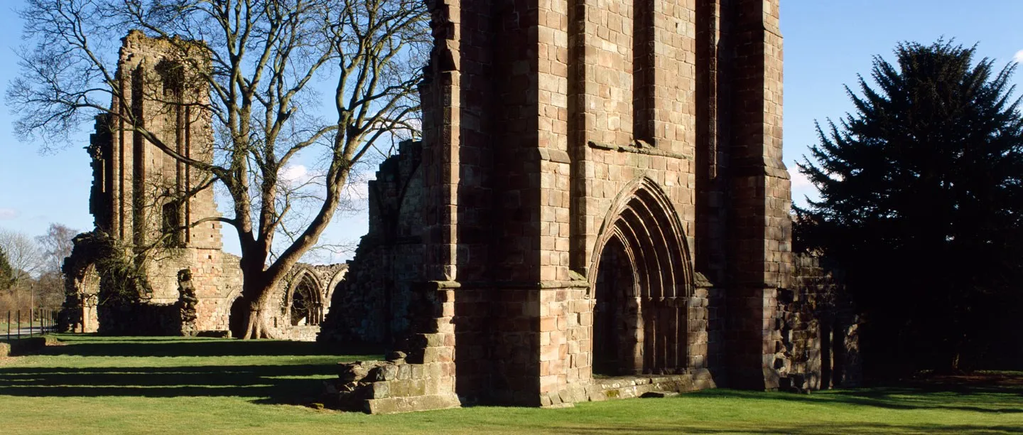 The tall west front of Croxden Abbey with its deeply moulded doorway, beside a bare tree, a dark yew and the transept ruins