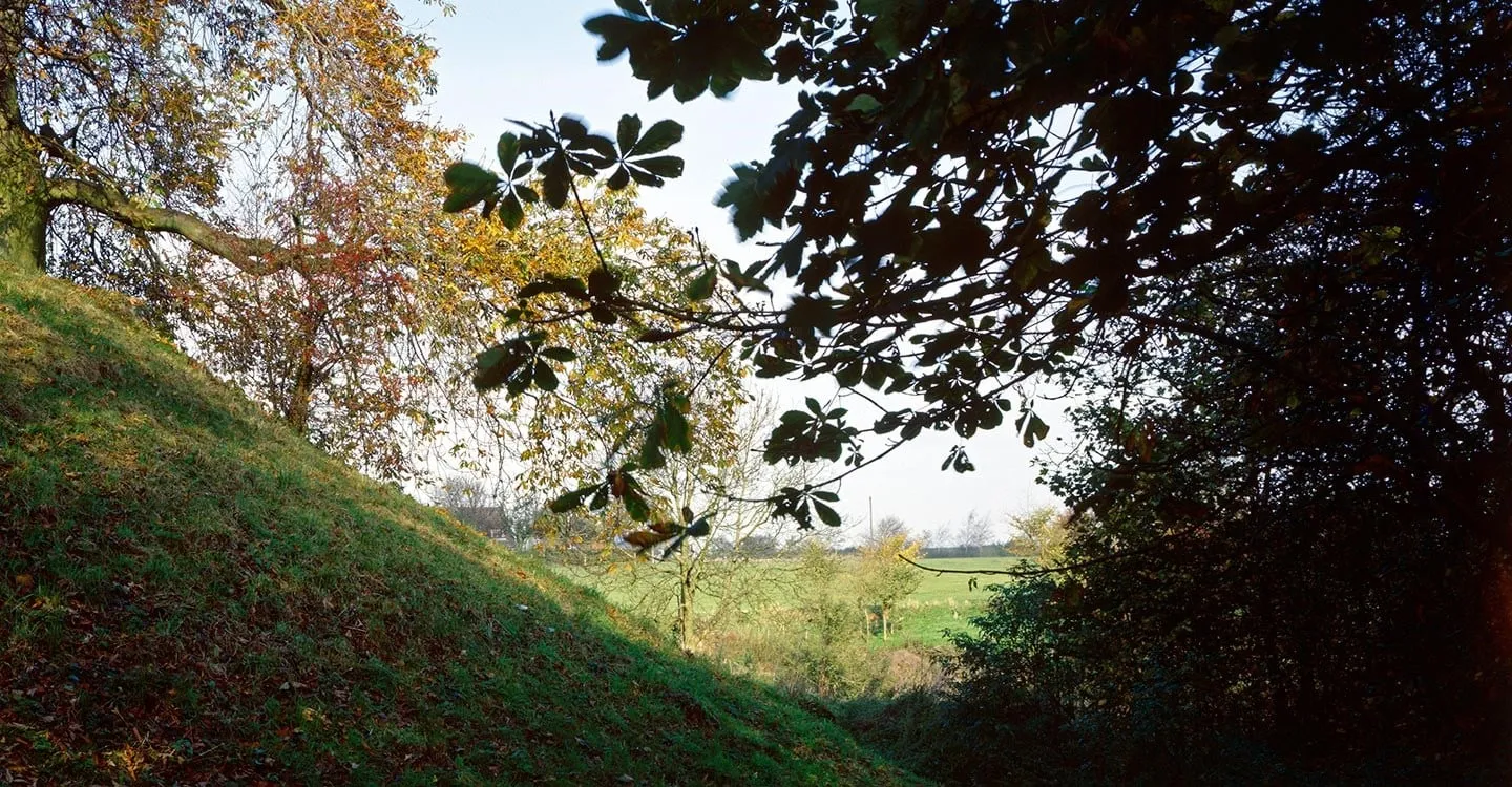The steep grassy earthwork bank of Deddington Castle under autumn horse chestnut trees, with fields beyond