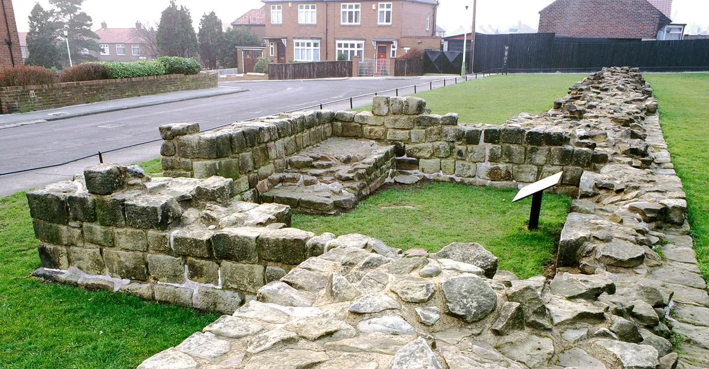 The squared stone walls of Denton Hall Turret and a stretch of Hadrian's Wall on a grass verge beside a suburban road