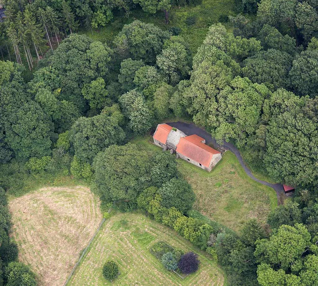 An aerial view of Derwentcote Steel Furnace's red roofs and chimney in a clearing in dense woodland