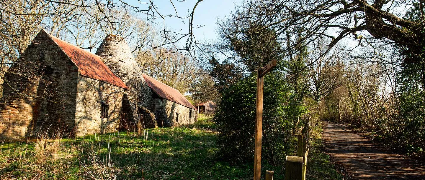 Derwentcote Steel Furnace beside a woodland path and wooden fingerpost, its tall conical chimney rising between two tiled roofs