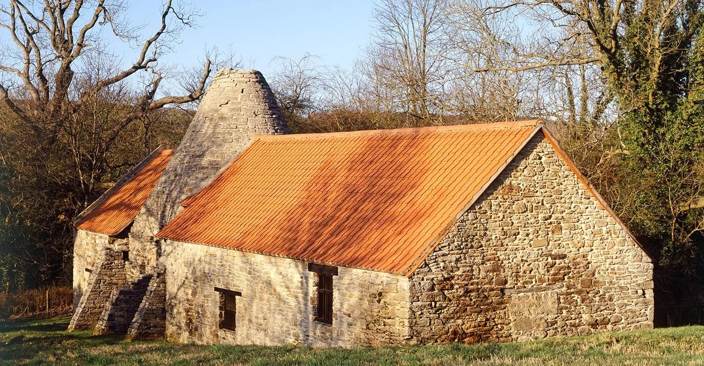 Derwentcote Steel Furnace, a stone building with a conical chimney and orange pantile roofs, in low winter sun among bare trees